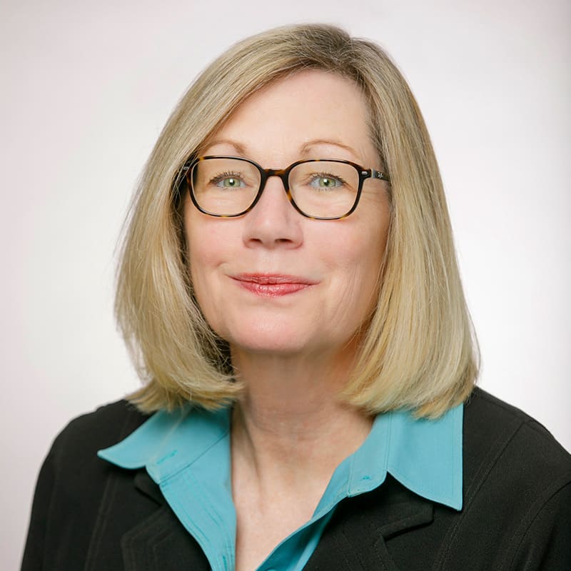 Portrait of Leeann Davis, a blonde woman wearing glasses and a black blazer over a turquoise blouse